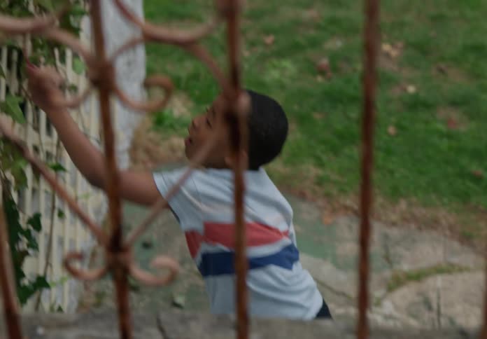 A young boy with short dark hair wearing a striped shirt plays by slapping a toy against a fence while an adult surprises him from a balcony