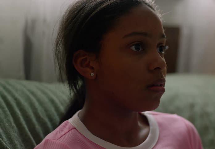 A young person with long dark hair pulled back and wearing a pink shirt looks off to the right with a focused expression
