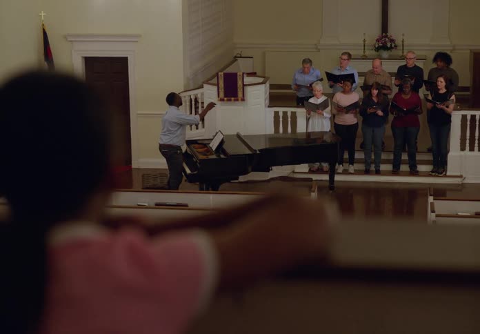 A black man plays a grand piano while a choir sings holding hymn books as a little girl in the audience watches