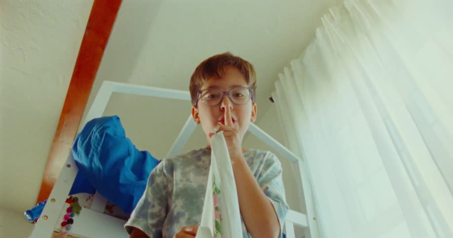 A boy looks at the camera as if its another child in a game of hide and seek signals for silence with a finger to his mouth glances around then covers the camera with a blanket as if hiding