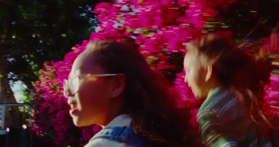 Two girls with flowing hair are captured in motion against a vibrant pink floral background in a sunny backyard