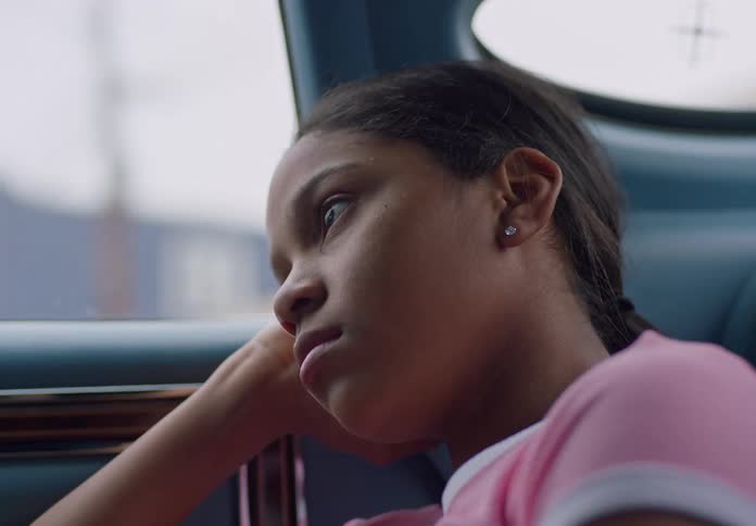 A teenage black girl sits in the back of a moving car at midday looking sad and bored