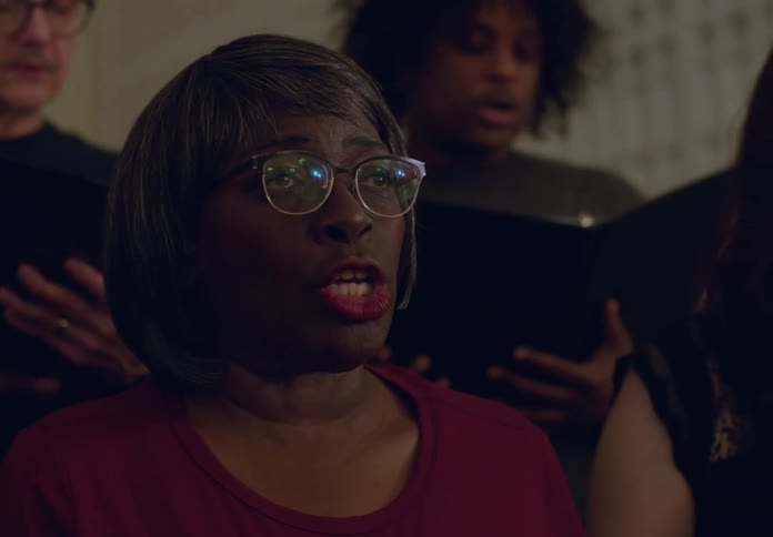 Several adults are singing together holding sheet music in what appears to be a choir practice