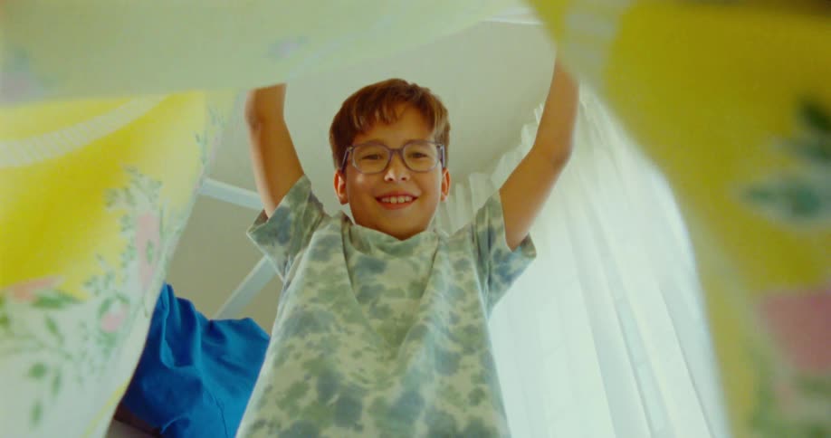 A young boy excitedly lifts a blanket indoors while playing hide and seek