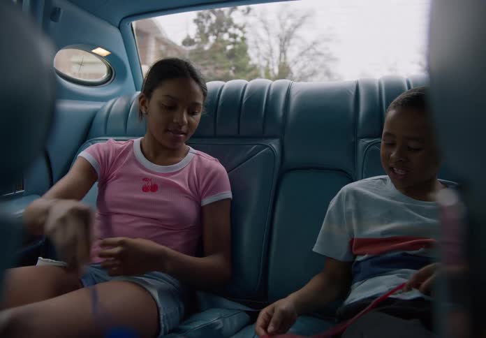 A boy and a girl sit in the back seat of a car at midday playfully interacting with each other