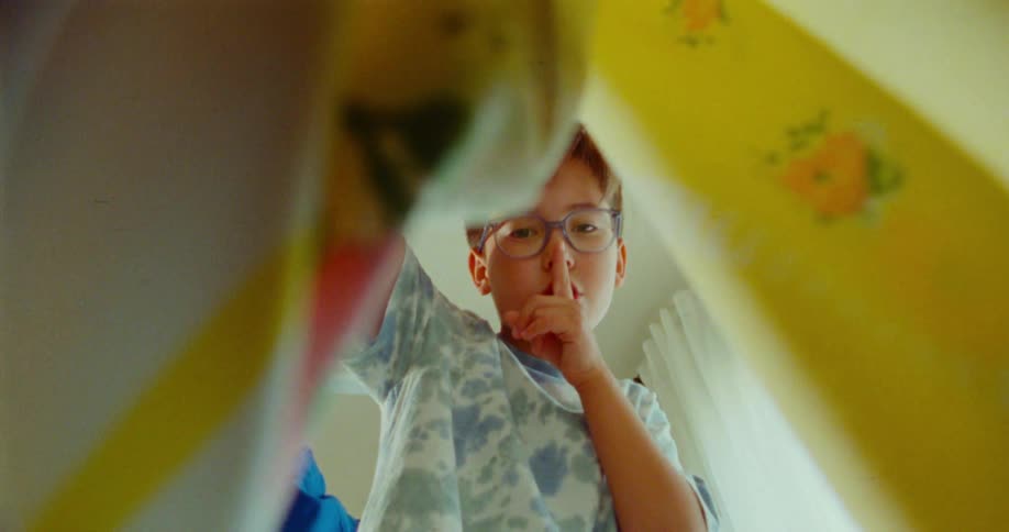 A child wearing glasses and a patterned shirt lifts a blanket looks down and places a finger to their lips as if requesting silence