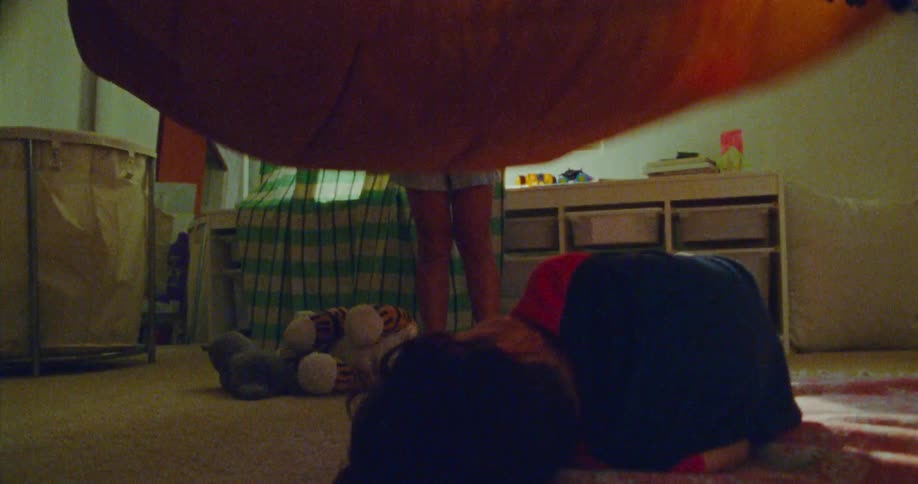 A group of children play together in a sunny room flapping a blanket as one child jumps under it and gets covered