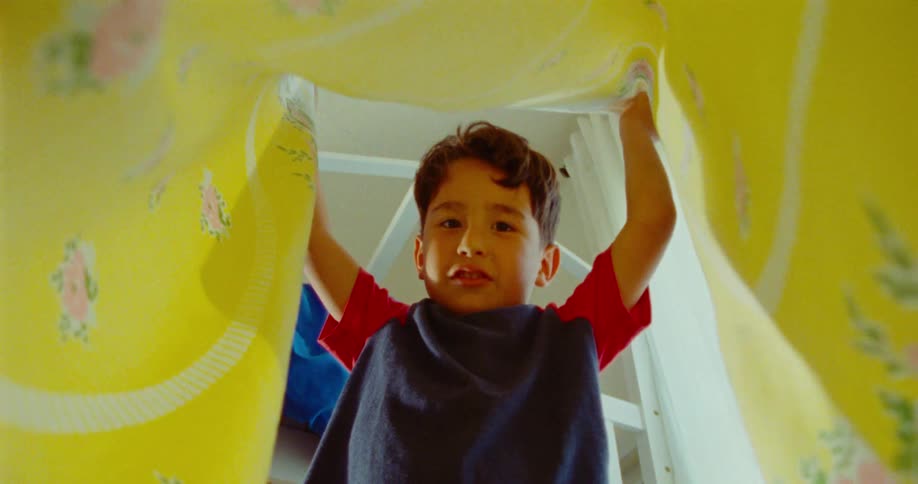 A young boy smiles playfully indoors while lifting a blanket
