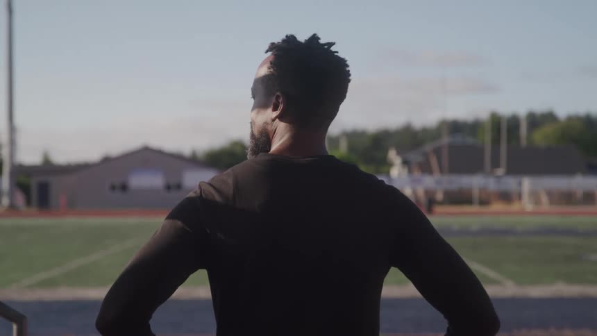 A person with dark skin and short textured hair wearing a dark longsleeved shirt stands with their hands on their hips facing a sports field with running tracks