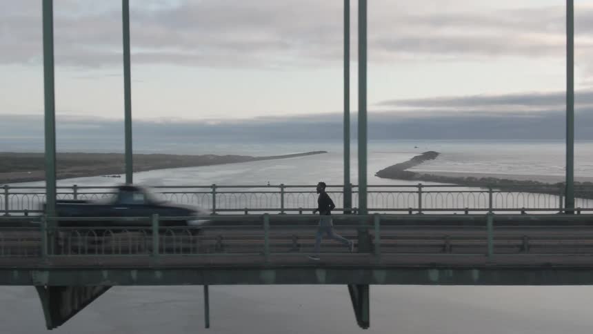 A person jogs along the pedestrian walkway of a large bridge spanning a body of water under an overcast sky
