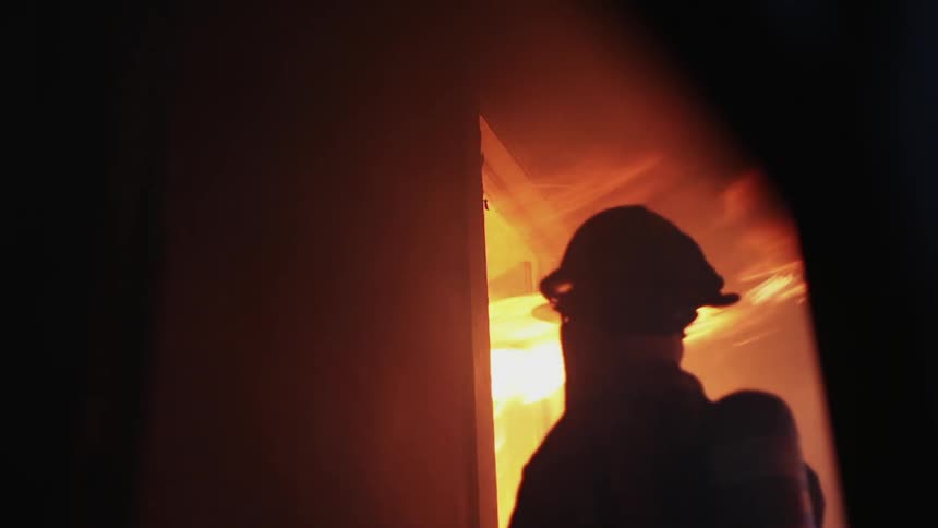 A firefighter is standing outside of a burning doorway
