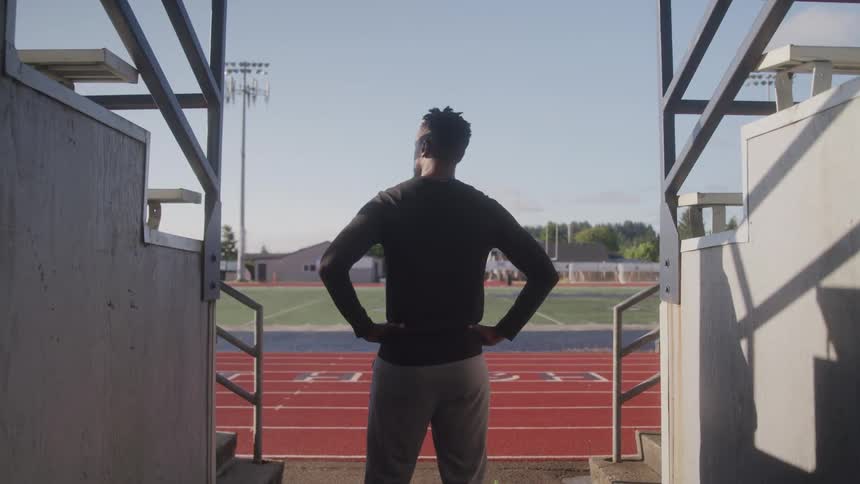 A black man stands at the entrance of a football field on a sunny day with both hands on his waist watching the field