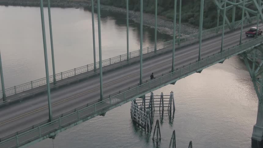 Aerial view of a person running alongside a bridge with cars passing on the street overhead and he is running over a body of water