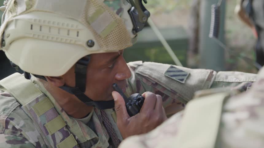 The face of a soldier wearing a helmet and military equipment and he is using a walkietalkie near his mouth to talk while pointing at something in front of him