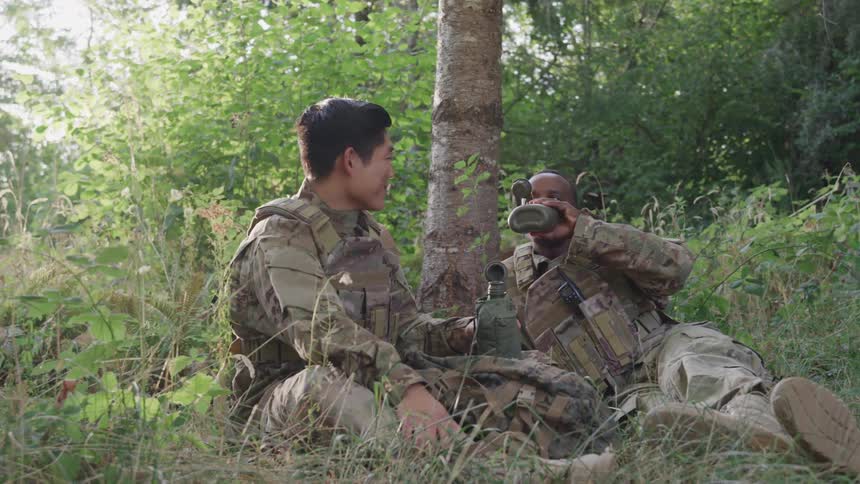 Two soldiers are sitting by the tree drinking from a canteen