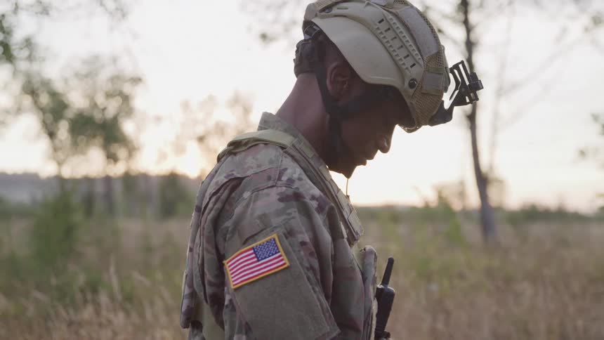 A military officer stands in the jungle at midday looking down while wearing a helmet and a walkietalkie attached to his front pouch