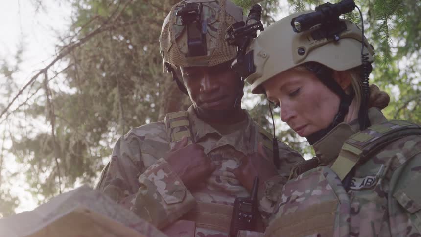 Two soldiers in camouflage uniforms and tactical gear are standing close together in a wooded area reading a map