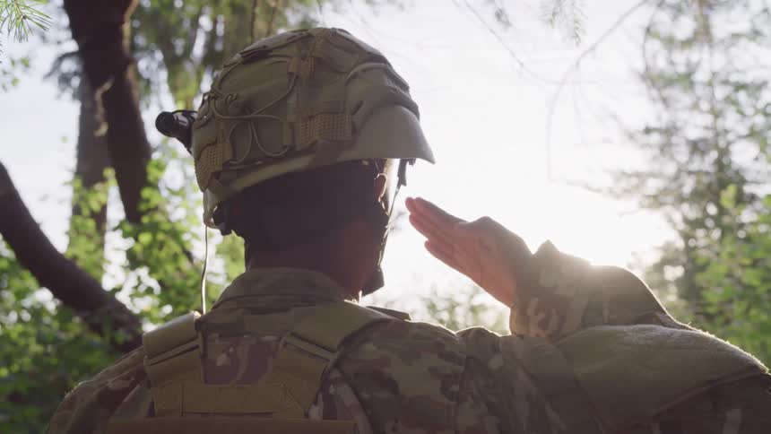 A soldier in camouflage uniform and helmet is saluting outdoors in a forested area with sunlight filtering through the trees