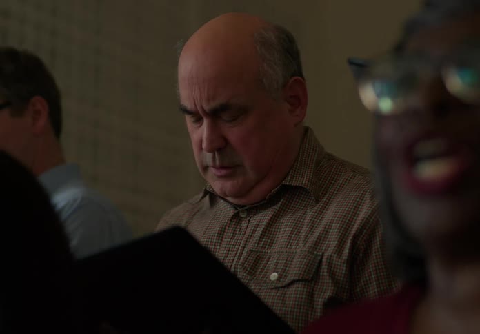 An older man is singing or reading in a choir setting