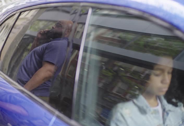 A person in a blue shirt is reflected in the window of a blue car talking on the phone while a girl sits inside the vehicle