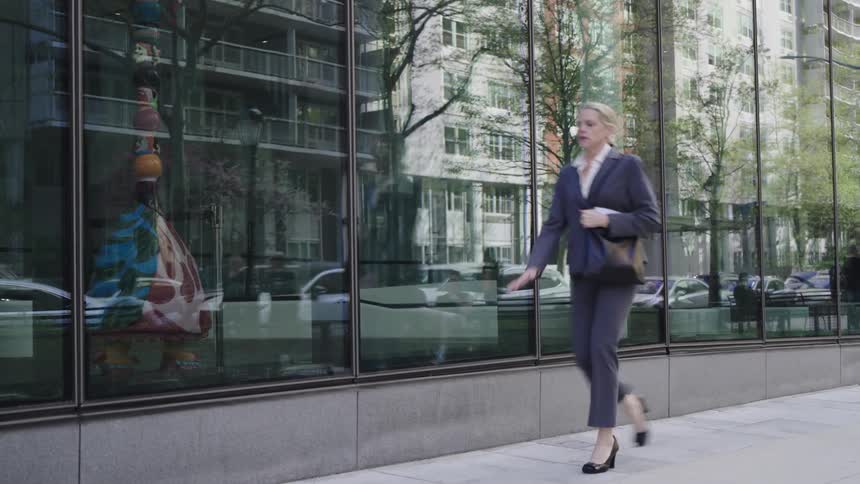 Woman wearing a pantsuit walks past a building with reflective glass during the daytime