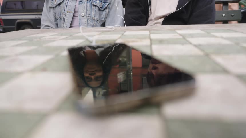 Smartphone reflects two teenagers sharing earbuds while pedestrians walk past