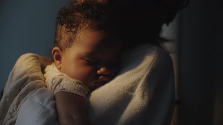 Woman holds a sleeping infant while standing in a dimly lit room