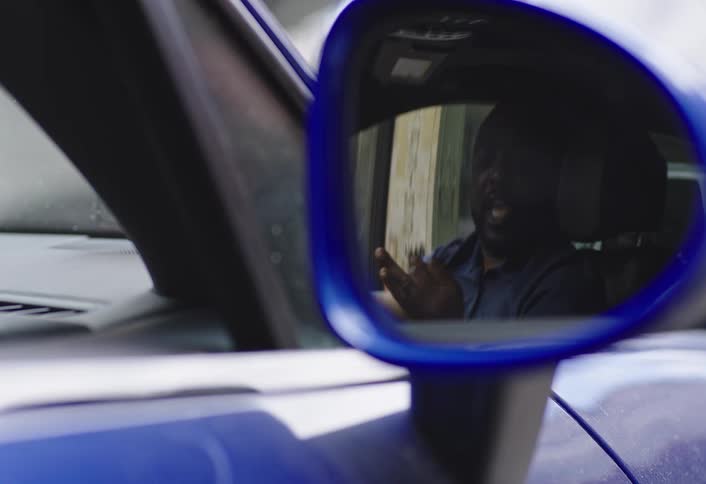 A man is seen through a cars side mirror sitting inside a blue car and talking to another person at midday