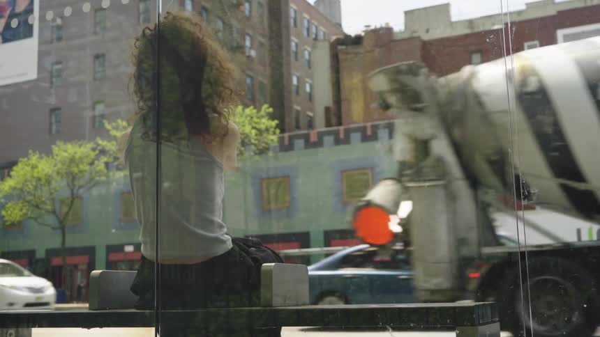 A woman sits inside by a glass window with city buildings and traffic reflected behind her