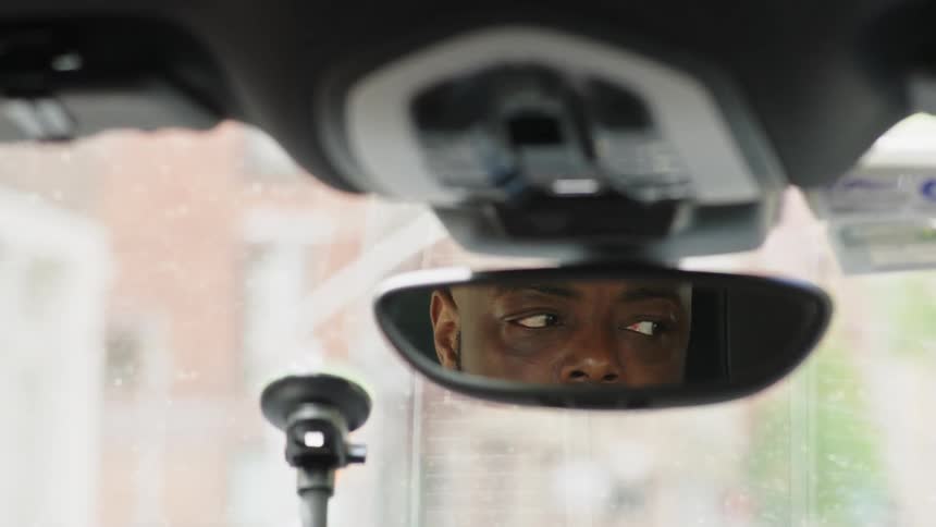 A young black man sits inside his car seen through the rearview mirror