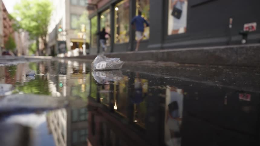 Two people run on the sidewalk near a discarded plastic bag floating on a reflective street puddle