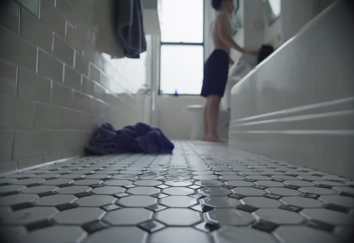 A person in shorts stands at a bathroom sink cleaning the mirror with a towel on the tiled floor