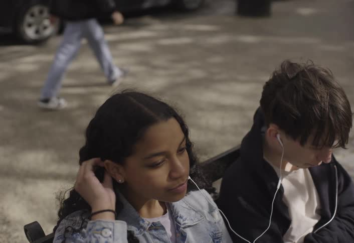 Two teenagers a boy and a girl sit on a bench at the side of the road at midday sharing a wired earbud and listening to a song