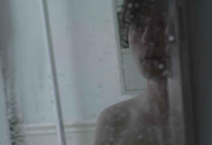 A male teenager stands in the bathroom staring at himself in the mirror