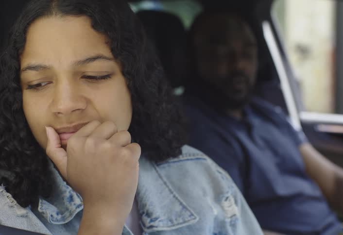 A black man scolds a teenage girl as they sit in the car at midday while the girl turns her face away from him looking sad
