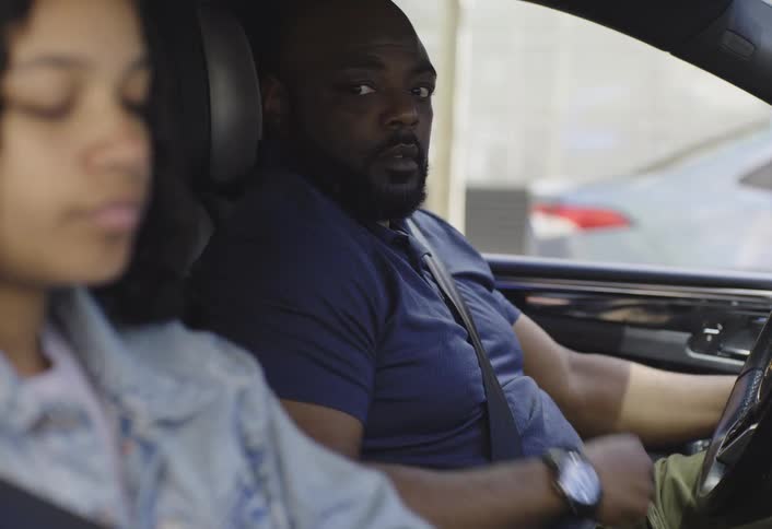 A man and a teenage girl inside a car put on their seat belts as the man prepares to drive