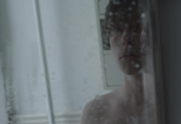 A shirtless person looks at their reflection in a foggy bathroom mirror