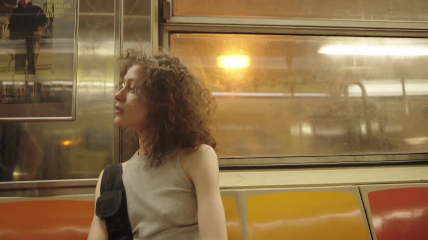 A young woman with curly hair and a serious expression sits on an orange and red subway seat gazing off to the side