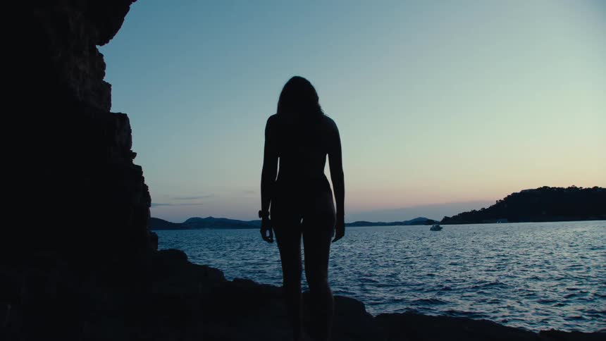 Silhouette of a woman in a bikini walking on a rock by the sea at dawn