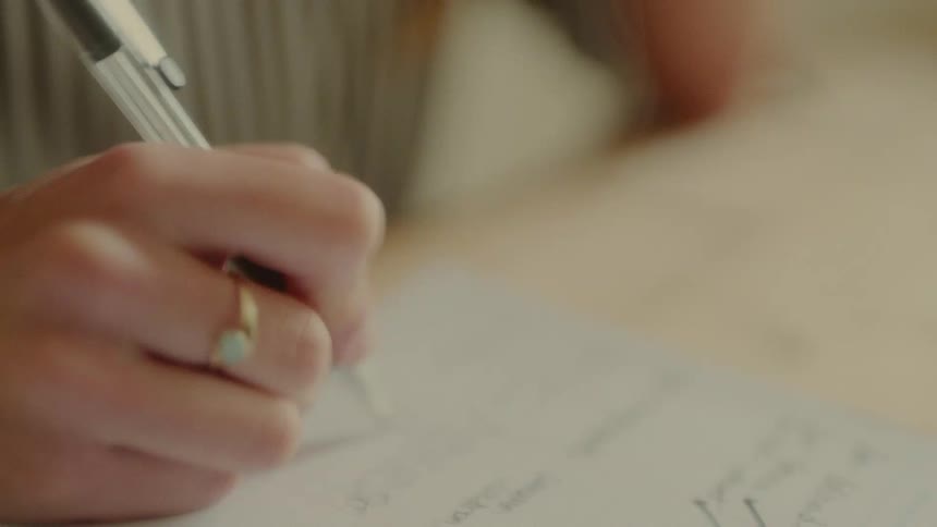 A hand with a bracelet and a ring holds a pen writing on a white piece of paper