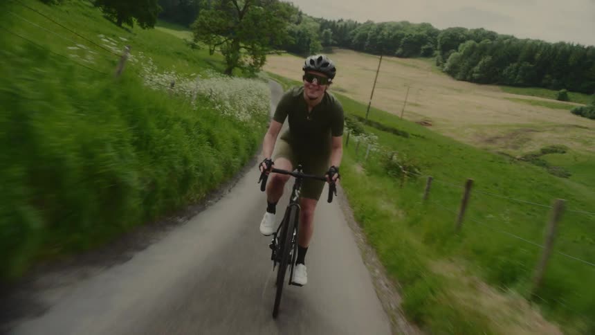 A person in a cycling kit helmet and sunglasses rides a bicycle down a winding road through a lush green rural landscape