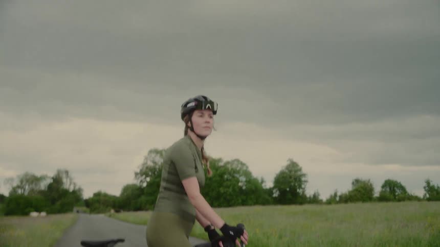 Female cyclist standing on a bicycle on a road surrounded by rural surroundings
