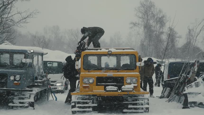 A man on top of a snow vehicle receives a snowboard from another man standing on the ground
