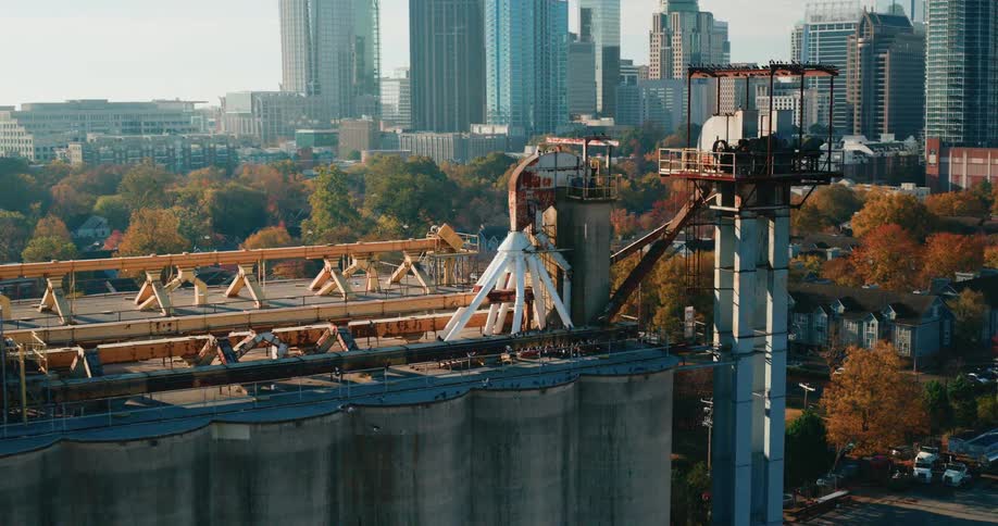 Large industrial silos stand in the foreground as birds circle above with a city skyline and autumn trees in the background