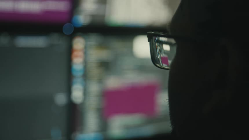 A person wearing glasses gazes at multiple blurry computer screens displaying various data and interfaces