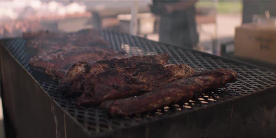 A barbecue stand grilling several steaks as a man walks back and forth nearby
