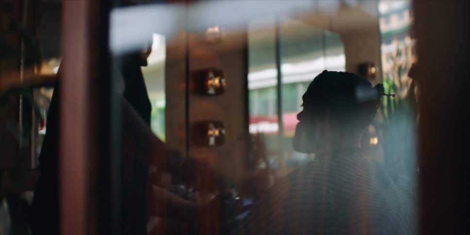 A barber cuts a clients hair inside a warmly lit shop viewed through a reflective glass window