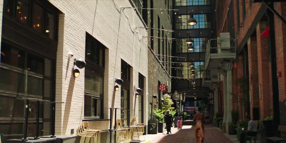 People walk down an alley leading towards a skyscraper with a reflective facade during a summer day