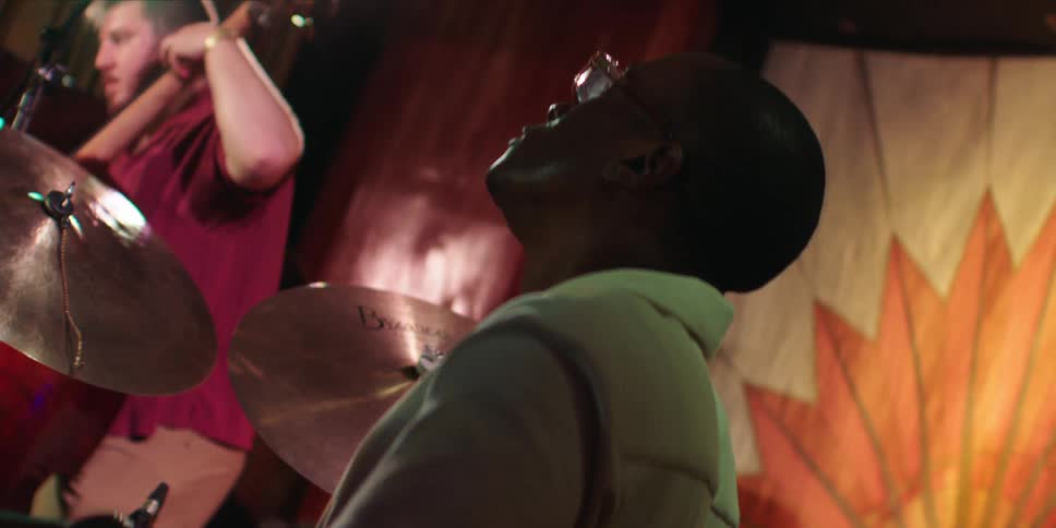 Man with glasses playing drums and in the background another person playing violin