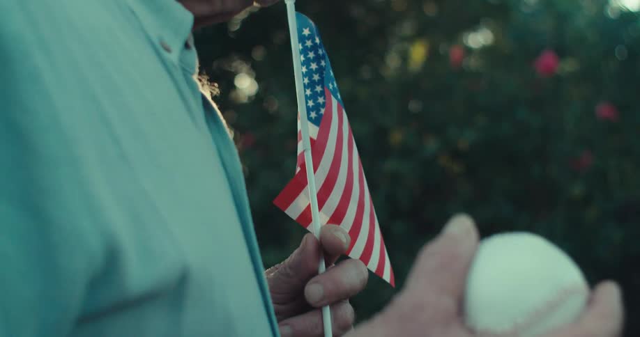 Elderly man standing outdoors holds a small american flag and a baseball